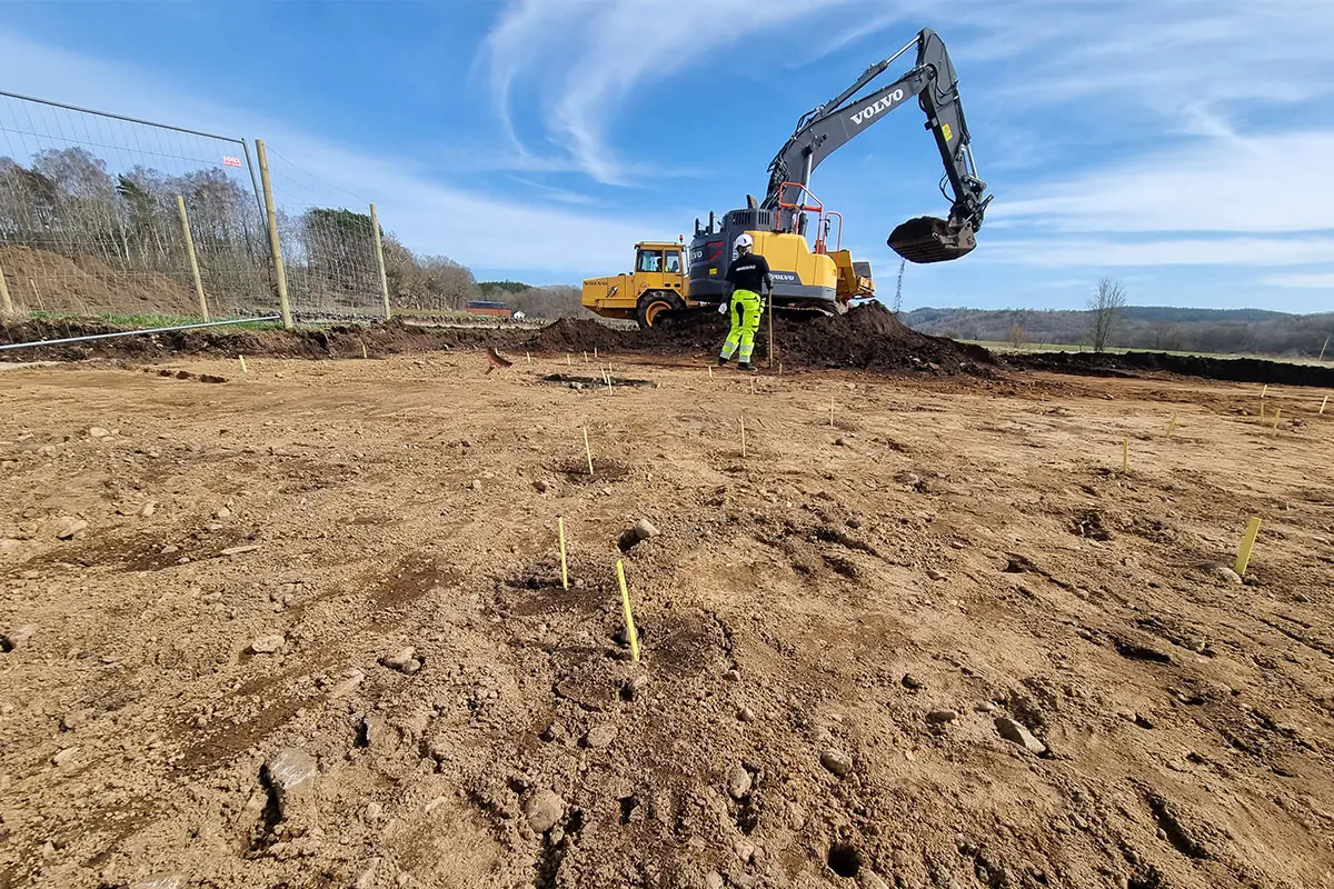 En stor yta med jord där arkeologer satt gula pinnar i marken. I bakgrunden står en grävmaskin och en person strax utanför den.